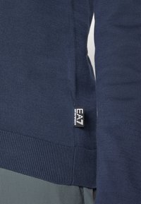 Mörkblå tröja som visas vid armen, med en ribbad nederkant och en EAN-etikett på sidan, tillverkad av mjukt stickat tyg.