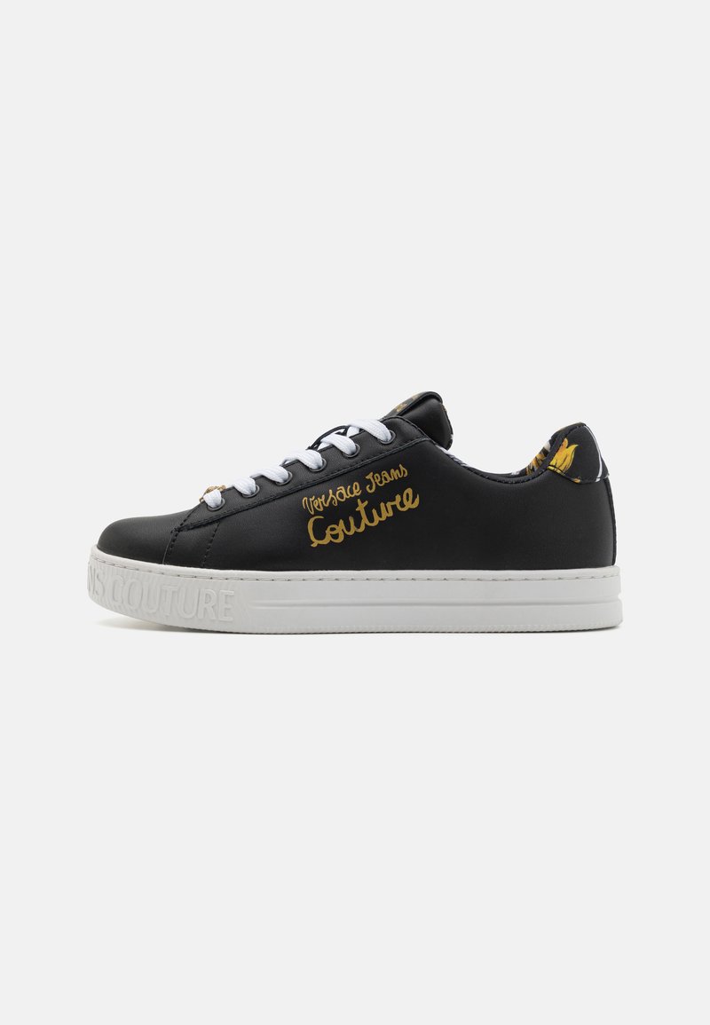 Sneaker en cuir noir avec semelle en caoutchouc blanche, comportant le texte doré "Versace Jeans Couture" et un design à lacets, agrémenté d'un talon à motif.