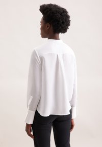 Camicia bianca a maniche lunghe con righe verticali, polsini con bottoni e piega sul retro, abbinata a jeans neri. Tessuto dalla texture liscia.