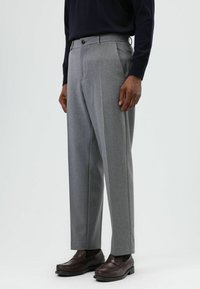 Grijze, op maat gemaakte broek met plooien, voorzien van een platte voorkant, riemlussen en twee zijzakken. Gecombineerd met donkerbruine leren schoenen.