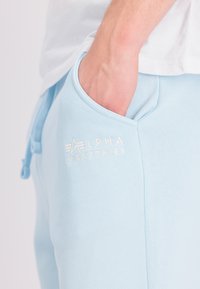 Svijetloplave trenirke s mekom teksturom, s bočnim džepovima i bijelim izvezenim logom "ALPHA INDUSTRIES" na butu.