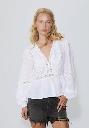 White long-sleeve blouse with a V-neck, intricate embroidery, and lace accents on the bodice. Paired with distressed black shorts.