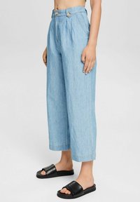 Femme portant un pantalon large bleu clair raccourci et des sandales noires à enfiler, debout devant un fond clair uni.