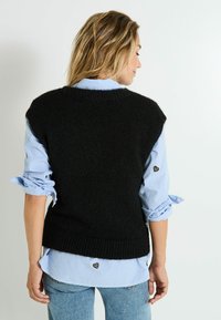 Gilet sans manches noir avec un design tricoté texturé, porté par-dessus une chemise à rayures bleues et blanches avec des accents en forme de cœur sur les poignets.