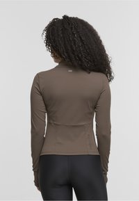 Brown long-sleeve top with a high neck, fitted design, and smooth texture. Features a logo detail at the back and is paired with black leggings.