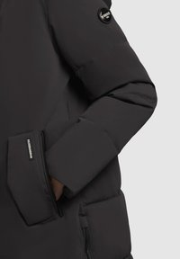 Zwarte pufferjack, met een gestructureerde afwerking, een ritszak en een rond logo-patch op de mouw.