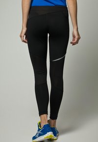 Black athletic leggings with a seamless design, featuring textured panels and reflective accents, paired with blue and yellow running shoes.