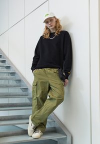 Schwarzer strukturierter übergroßer Pullover, olivgrüne Cargo-Hose mit Taschen, weiße Kappe mit Logo, beige Sneaker mit grauen Akzenten, silberne Halskette.
