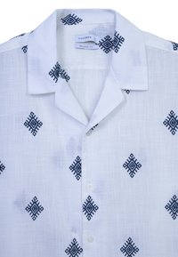 Camicia a maniche corte, dal taglio comodo, in tessuto bianco con un motivo geometrico nero. Dettaglio del colletto e della patta con cinque bottoni visibili.