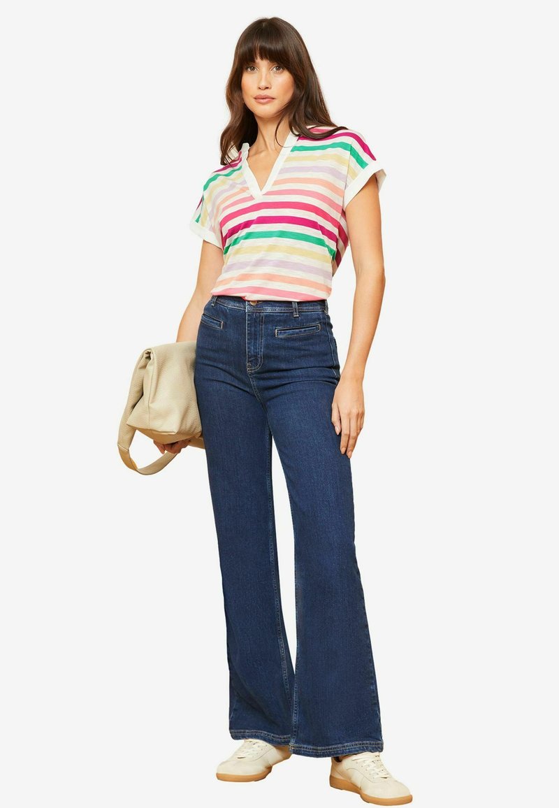 Rausvų, žalių ir smėlio spalvos dryžuotas trumpomis rankovėmis viršus, derinamas su tamsiai mėlynomis aukštu juosmeniu plačiomis džinsomis ir baltais sportbačiais, laikantis smėlio spalvos rankinę.