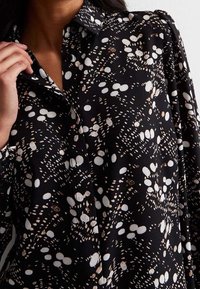 Musta pusero, jossa on valkoisia ja beigejä pisteitä eri kokoisina, klassinen kaulus ja napitus edessä. Tasainen, kevyt kangas.