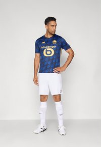 Futbalový dres modro-čierny s geometrickým vzorom a žltým logom, v kombinácii s bielymi šortkami a ponožkami a čiernymi kopačkami.