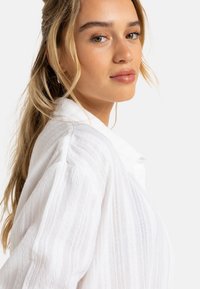White textured shirt with vertical stripes, featuring a classic collar and buttoned front, displayed on a person against a neutral background.