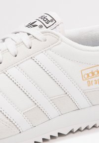 Weiße Adidas Sneaker mit Obermaterial aus Wildleder und Canvas, ausgestattet mit drei charakteristischen Streifen, strukturierten Details und einer strukturierten Sohle.