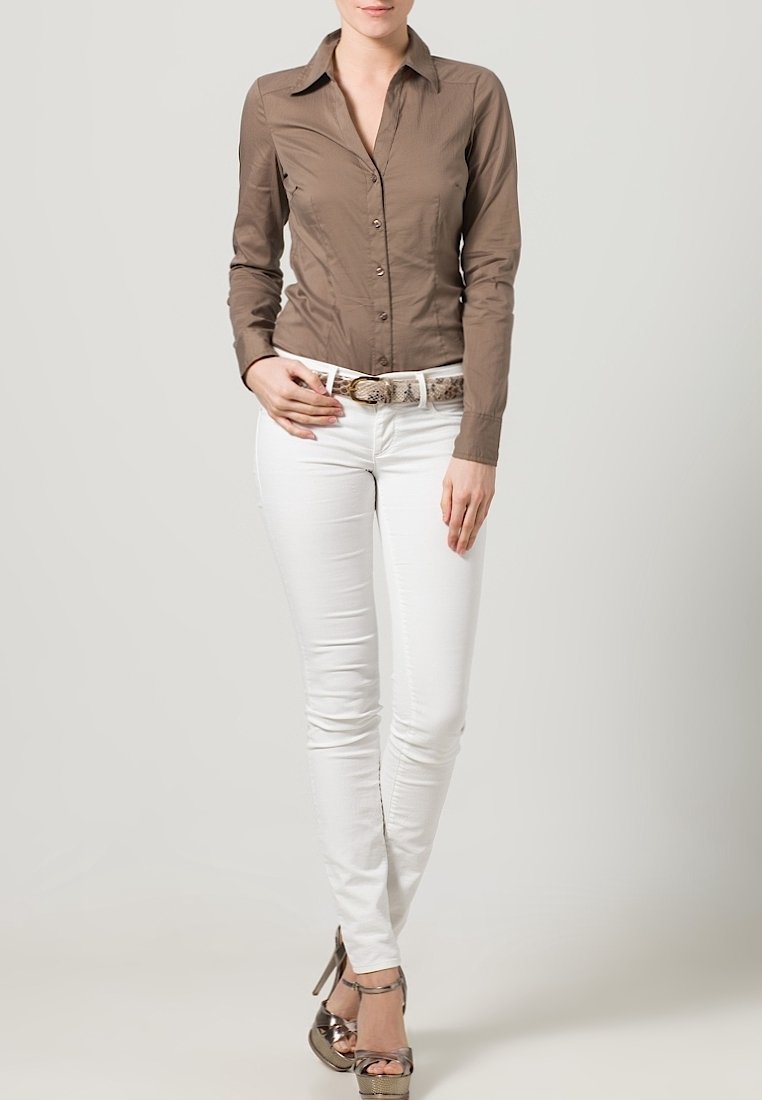 Femme portant une chemise marron cintrée à boutons, un pantalon blanc avec une ceinture à motifs et des sandales métalliques à talons hauts, debout sur un fond uni.