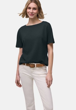 Kvinde med kort blondt hår iført en sort tekstureret kortærmet top, beige bukser og et brunt bælte, stående mod en ensfarvet baggrund.