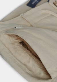 Gros plan d'une poche de vêtement en lin beige avec une fermeture éclair en métal marron visible, partiellement ouverte.