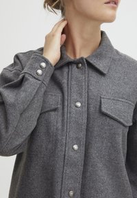 Szara koszula-kurtka z mieszanki wełny z ostrym kołnierzem, zapięciem na zatrzaski, dwoma przednimi kieszeniami oraz teksturowaną powierzchnią tkaniny.