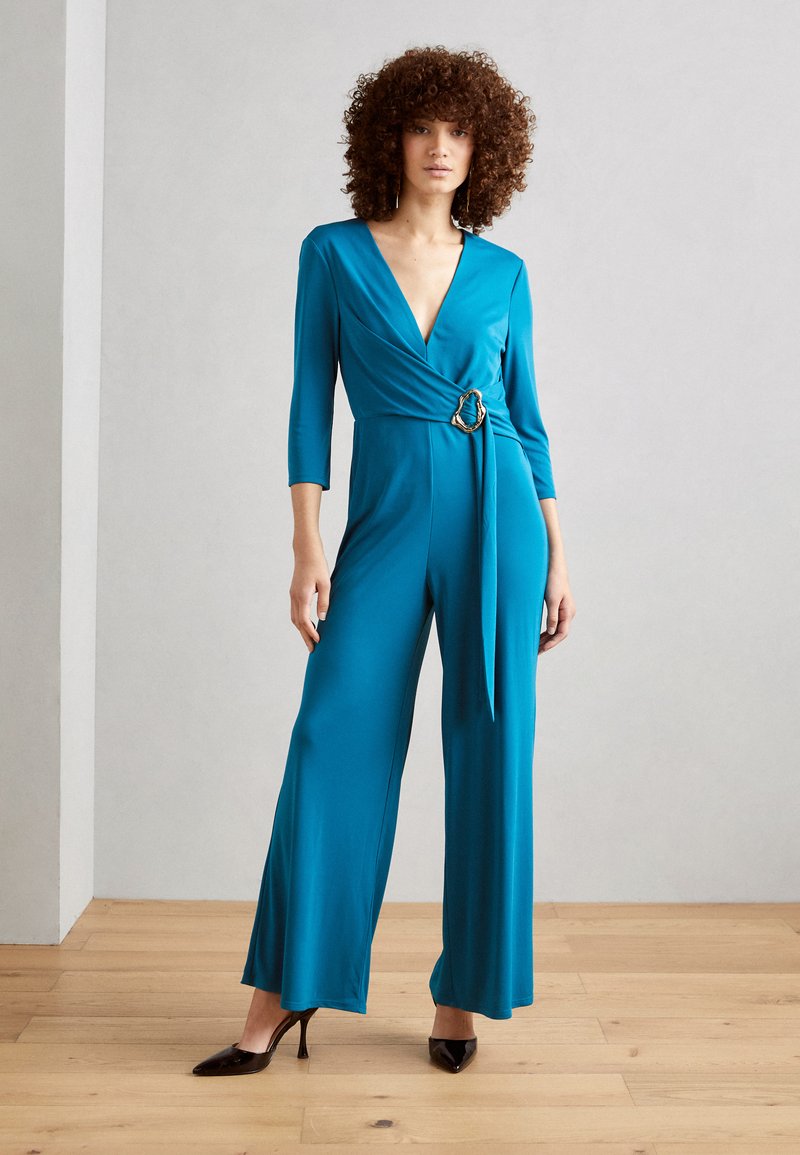 Teal jumpsuit med dyp V-hals, trekvart erm og midjebånd med en rund detalj i gullring. Glatt, elastisk stoff, vide ben.