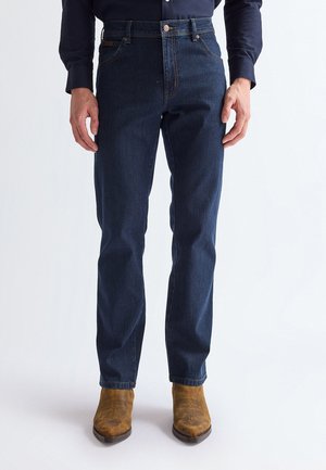 Ein Mann trägt dunkelblaue Jeans mit geradem Bein, ein schwarzes Hemd und braune Wildleder-Westernstiefel und steht vor einem schlichten Hintergrund.