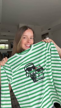 Junge Frau lächelt und hält ein grün-weiß gestreiftes Hemd mit Cartoon-Figuren, die Sonnenbrillen tragen.