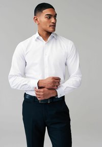 Camisa blanca de manga larga con una textura sutil, botonadura en el frente y pantalones de vestir negros en contraste, complementados con un cinturón oscuro.