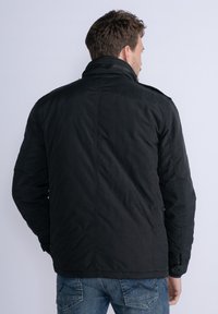 Giacca nera imbottita con colletto alto, cuciture a vista e dettagli in bottoni. Indossata con jeans di denim, presentando una vista posteriore semplice.