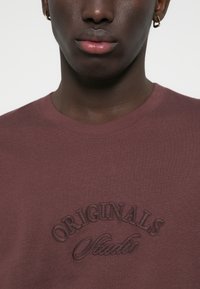 Bruine katoenen t-shirt met een gestructureerd, geborduurd "ORIGINALS Studio" logo op de borst. Eenvoudige ronde halslijn en korte mouwen.