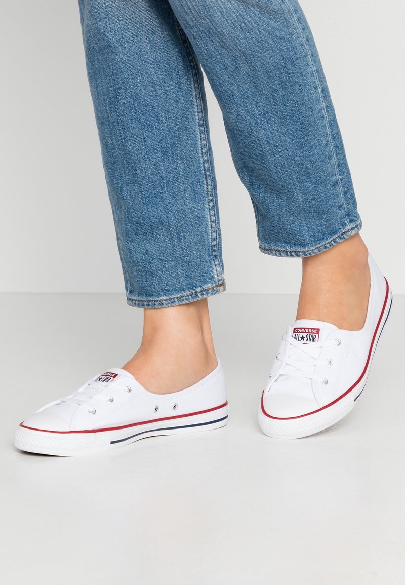 Jambes portant un jean bleu et des baskets basses Converse blanches avec un détail de rayures rouges et bleu marine sur les semelles.