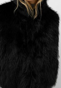 Musta tekoturkisliivi, jossa on pehmeä, pitkäkarvainen rakenne ja korkea kaulus. Suunnittelu on hihaton, mikä korostaa ylellistä materiaalia.