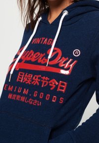 Sudadera azul marino con cordones blancos, que presenta un gran texto en rojo "Super Dry" y caracteres japoneses en la parte delantera.
