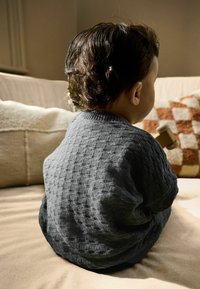 Gray textured sweater with a ribbed neckline, worn by a child holding a wooden toy, seated against a light-colored backdrop.