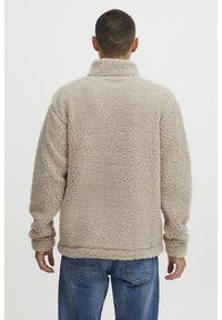 Polarowy sweter w jasnym beżu, z wysokim kołnierzem, długimi rękawami i teksturowaną powierzchnią. Żebrowane wykończenia u dołu i na mankietach.