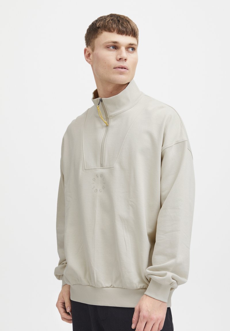 Sudadera de medio cremallera color beige claro con un cuello alto, puños y dobladillo elásticos, que presenta un detalle de logo texturizado en el frente. Textura de tela suave.