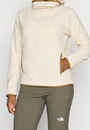Persoon draagt een crème fleece trui met bruine afwerking en groene North Face broek, hand in de trui-zak tegen een effen achtergrond.