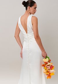 Femme aux cheveux foncés en chignon portant une robe blanche dos nu avec des boutons, tenant un bouquet de fleurs orange, jaune et rose, vue de profil.