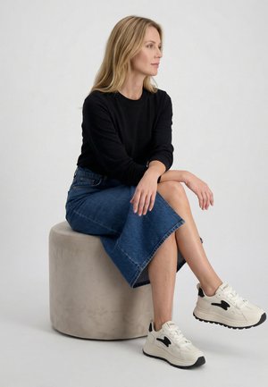 Kobieta o blond włosach, ubrana w czarną bluzkę z długim rękawem, niebieską dżinsową spódnicę i białe trampki, siedząca bokiem na okrągłym beżowym stołku.