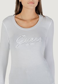 Top de manga larga blanco hecho de una tela suave, con un escote redondeado y un logotipo "Guess" adornado con detalles en plata.