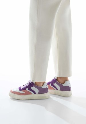 Sneakers feature a white leather base with violet and pink suede panels, textured rubber sole, and round laces. Minimalist design with accents.