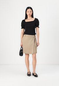 Top nero a coste con maniche a sbuffo corte abbinato a una gonna avvolgente beige. La modella tiene in mano una piccola borsa nera e indossa scarpe nere basse.