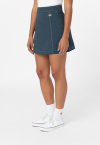 Tmavě tyrkysová sukně s bílým prošitím, A-liniový střih a malý detail loga na přední straně. Nosená s bílými ponožkami a botami.