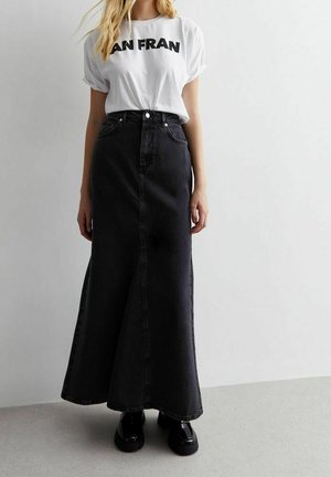 Vrouw die een wit T-shirt met opgerolde mouwen draagt met de tekst "SAN FRAN" en een zwarte denim maxi-rok met hoge taille en zwarte schoenen, staand binnen.