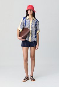 Camicia a maniche corte con stampa paisley blu e gialla, pantaloni corti blu scuri, cappello a secchiello rosso, pochette marrone e sandali neri con dettagli dorati.