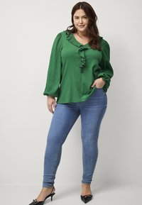 Grøn tekstureret bluse med rynket halsudskæring, parret med tætsiddende blå jeans og sorte spidse sko.