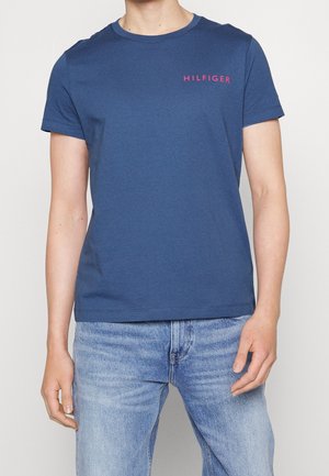 Mężczyzna ubrany w dopasowaną granatową koszulkę z czerwonym napisem „HILFIGER” na piersi oraz jasne niebieskie dżinsy.