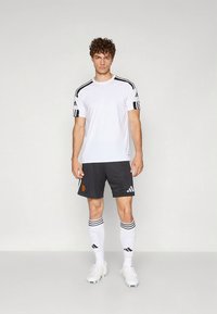 Baltas drėgmę sugeriantis džersis su juodomis juostomis, derinamas su tamsiai pilkomis šortais su logotipu, baltos kojinės su juodomis juostomis ir futbolo bateliai.