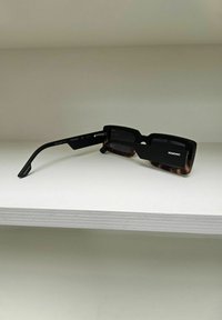 Lunettes de soleil noires avec une monture rectangulaire, présentant un extérieur texturé et des accents en écaille de tortue sur les branches. Nom de la marque "Komono" visible.