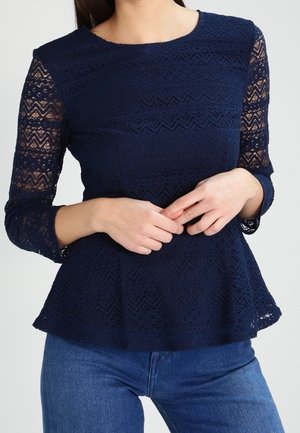 Vrouw draagt een marineblauw kanten peplumtop met driekwartmouwen en blauwe jeans, handen losjes gevouwen ter hoogte van de taille.