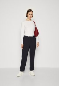 Maglietta bianca a maniche lunghe, pantaloni navy con cintura "HILFIGER", sneakers bianche e una borsa a tracolla rossa. Tessuti lisci e vestibilità sartoriale.
