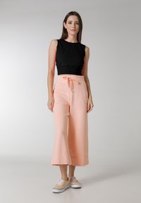 Zwarte mouwloze top met geplooide details, gecombineerd met lichtroze wijde broeklegs met een elastische tailleband en trekkoord. Beige sneakers.